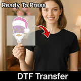 A smiling woman in a black t-shirt holds an InkMerge "Skull with Santa Hat Dark Humor Holiday" DTF transfer sheet, showing a colorful and humorous holiday design. The image includes "Ready To Press" and "heat press transfer" text.