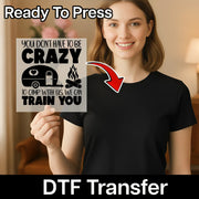 A smiling woman in a black shirt holds InkMerge’s Crazy Camper Humor DTF transfer, featuring a camper, campfire, and the quote: “You don't have to be crazy to camp with us, we can train you.” Perfect for outdoor adventure apparel.