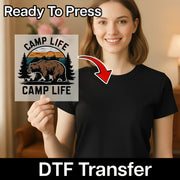 A smiling woman in a black shirt holds InkMerge’s Bear Mountain Sunset Design DTF transfer, featuring a bear, trees, and sunset. Text reads “Ready To Press” and “DTF Transfer”—ideal for custom apparel or any heat press project.