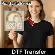 A smiling woman in a black shirt holds the InkMerge Be Kind Retro Rainbow transfer, featuring bold retro colors and an uplifting design. Ready To Press and DTF transfers make this custom apparel piece easy to use and vibrant.