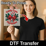 A smiling woman in a black shirt holds InkMerge's Candy Cane Christmas Design DTF transfer, featuring a candy cane, bow, and holly leaves. Ideal for holiday heat press projects! Text reads "Ready To Press" and "DTF Transfer.