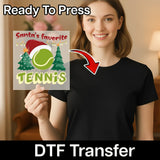 A smiling woman in a black shirt holds the Santa's Favorite Tennis DTF transfer by InkMerge, featuring a festive tennis design with Santa hat, trees, and tennis ball—perfect for game day outfits! "Ready To Press" appears above.