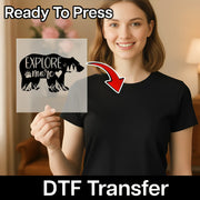 A smiling woman holds InkMerge's Explore More Bear Adventure Design—a clear DTF transfer featuring a black bear and "Explore more." An arrow points to her plain black tee. Text: Ready To Press DTF Transfer for heat press application.