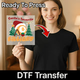 A smiling woman in a black shirt holds an InkMerge "Santa Billiard Humor" transfer featuring a Santa hat, billiard balls, trees, and an arrow—fun for your next ugly sweater party.