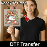A smiling woman in a black t-shirt holds an InkMerge Cowboy Christmas Snowman Design DTF transfer sheet featuring a snowman in a cowboy hat, cacti, and festive decor. Text: Ready To Press and holiday heat press transfers.