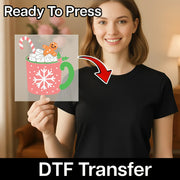 A smiling woman in a black shirt holds an InkMerge "Festive Gingerbread Mug Design - Heat Press Ready" DTF transfer, featuring marshmallows, candy cane, and gingerbread art. Text reads "Ready To Press"—perfect for your heat press!.