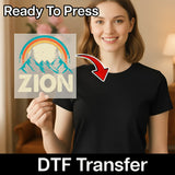 A smiling woman in a black shirt holds InkMerge’s Take a Hike Retro Mountain Design DTF transfer sheet with "ZION." “Ready To Press” and “DTF Transfer” text highlights its easy heat press application, with an arrow pointing to the shirt.