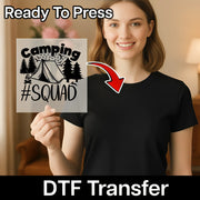 A woman in a black shirt holds an InkMerge Camping #Squad Outdoor Adventure - Heat Press Ready transfer with tent and trees design. "Ready To Press" appears above, "DTF Transfer" below, with an arrow from the transfer to her shirt.
