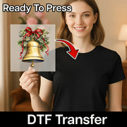 A smiling woman in a black shirt holds an InkMerge Golden Bell with Holly Design DTF transfer featuring a red ribbon and greenery. “Ready To Press” is at the top, “DTF Transfer” at the bottom—a great custom apparel screen printing alternative.