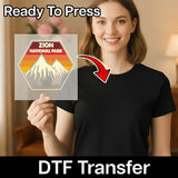 A smiling woman in a black t-shirt holds InkMerge's Zion National Park Retro Mountain Design - Heat Press Ready, featuring mountains and a sunset with "Ready To Press" at the top and "DTF Transfer" at the bottom—ideal for custom apparel.