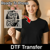 A smiling woman in a black shirt holds a transparent Camp More Worky Less Design sheet by InkMerge—ideal for custom apparel. An arrow points to her shirt. Text reads Ready To Press and DTF transfers.