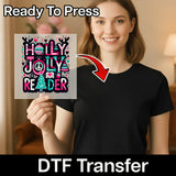 A smiling woman in a black shirt holds the InkMerge "Holly Jolly Reader Design" DTF transfer with festive Christmas motifs. An arrow points to her shirt. Text reads "Ready To Press" and "holiday heat press DTF Transfer.