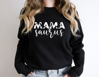 A woman with long, wavy hair (neck down) wears the InkMerge Mama Saurus black sweatshirt—ideal for mom hoodie fans. “MAMA” is printed in bold letters filled with dinosaur silhouettes.