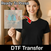 A smiling woman in a black shirt holds an InkMerge Warm Winter Wishes Vintage Design DTF transfer sheet featuring colorful winter trees. An arrow points to her shirt. Text reads Ready To Press and heat press transfer.