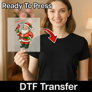 A woman holds a transparent InkMerge "A Little Christmas Miracle Santa & Cardinal" DTF transfer, featuring Santa and a cardinal, with an arrow to her plain black shirt. Labeled Ready To Press and Christmas DTF transfer.