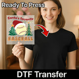 A smiling woman in a black shirt holds an InkMerge "Santa's Favorite Baseball Design" DTF transfer, featuring a baseball, Christmas tree, and Santa hat. An arrow points to her shirt—perfect for custom apparel and ready to press.