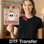 A smiling woman in a black shirt holds InkMerge's "Tis The Season To Be Jolly Santa Humor" DTF transfer, which is "Ready To Press." An arrow shows the festive holiday heat press design matching her shirt.