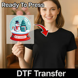A smiling woman in a black shirt holds an InkMerge Merry Bookmas Snow Globe Design, featuring a snowman and books. Labeled “Ready To Press” and “DTF Transfer,” it's the perfect festive touch for any ugly sweater party.