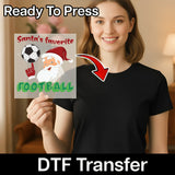 A smiling woman in a black shirt holds InkMerge's Santa's Favorite Football Soccer Design DTF transfer, featuring cartoon Santa and soccer ball, with "Ready To Press" above and "DTF Transfer" below—perfect for custom heat press projects.