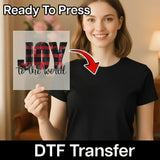 A woman in a black t-shirt holds an InkMerge JOY to the World Buffalo Plaid Design DTF transfer sheet. An arrow points to her shirt with text: Ready To Press DTF Transfer—compatible with any heat press.