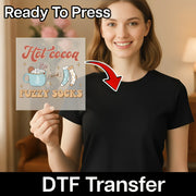 A smiling woman in a black t-shirt holds the InkMerge “Hot Cocoa Fuzzy Socks Winter Design” Ready To Press DTF transfer, with an arrow pointing to her shirt—perfect for your next heat press project.