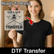 A smiling woman in a black shirt holds the InkMerge "Let's Get TOASTED Campfire Humor" DTF Transfer, which features marshmallows over a campfire and reads "Lets get TOASTED." Ready To Press—perfect for DTF transfers or custom apparel.
