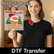 A smiling woman in a black shirt holds an InkMerge "Santa's Favorite Croquet" DTF transfer sheet with festive Christmas trees and a red hat. An arrow shows how easy it is to apply the design using a heat press.