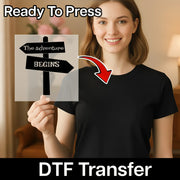 A woman in a black shirt smiles while holding InkMerge’s The Adventure Begins Signpost - Heat Press Ready DTF transfer, featuring a directional sign graphic—ideal for custom apparel with your heat press.